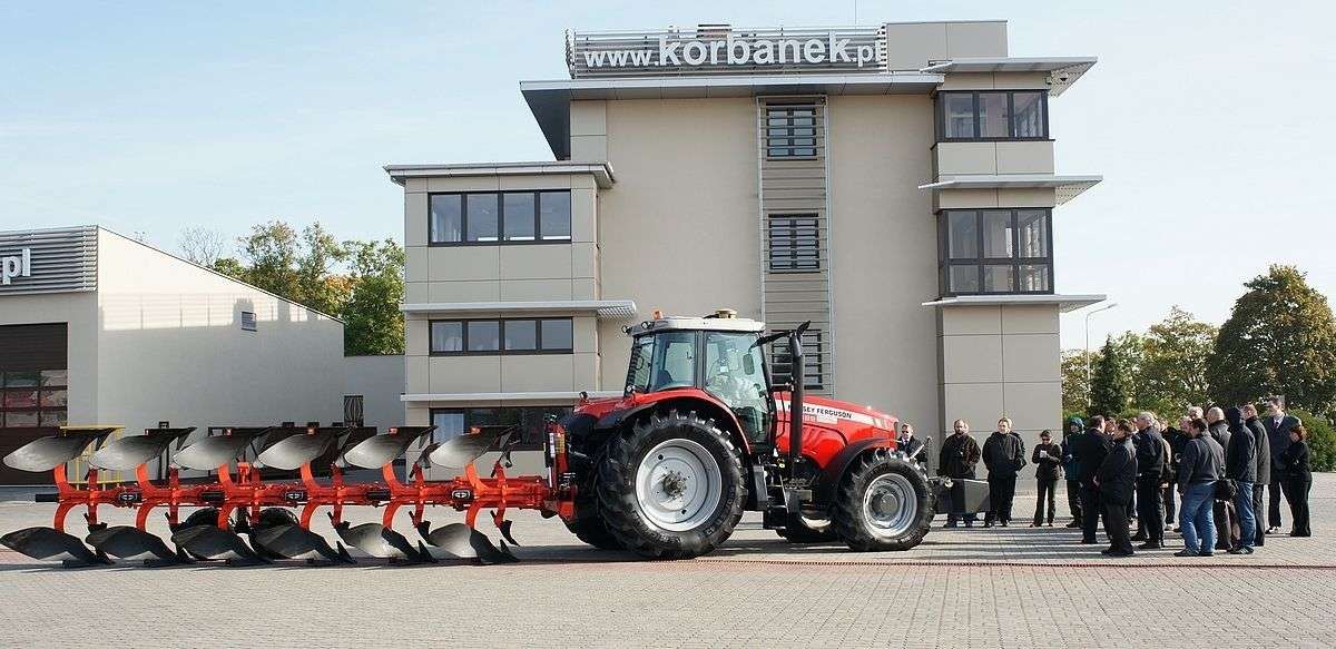 Massey Ferguson z pługiem obrotowym Gregoire Besson przed firmą korbanek ciągnik rolniczy Massey Ferguson z pługiem obrotowym przed firmą korbanek przed nim grupa zainteresowanych osób
